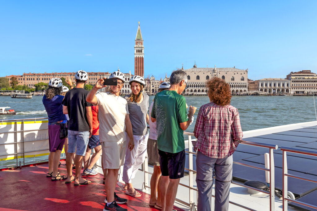 Gruppe von Touristen, die das Stadtbild von einem Boot aus betrachten, mit einem Glockenturm und historischen Gebäuden im Hintergrund.